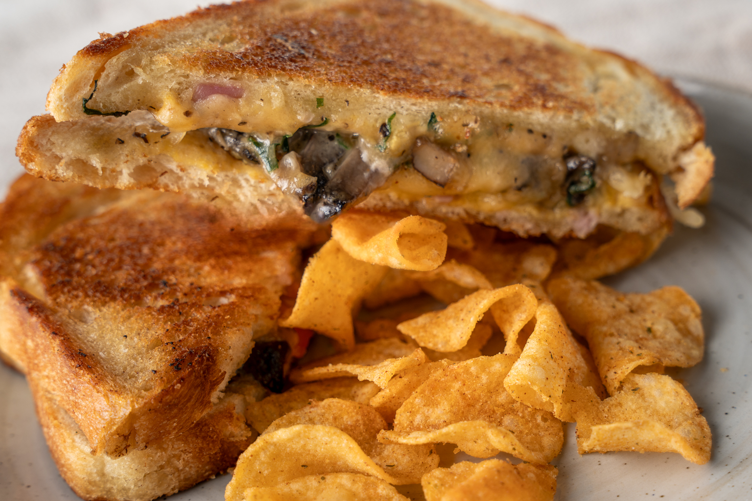 A close-up of a grilled cheese sandwich, featuring golden-brown toasted bread with melted cheese, mushrooms, and herbs, accompanied by a side of crispy potato chips on a white plate.