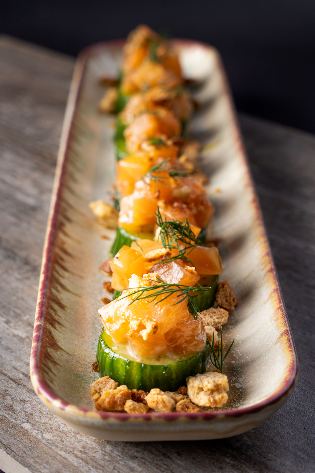 A beautifully arranged dish featuring cucumber slices topped with smoked salmon, dill, and crispy elements, served on an elegant platter.