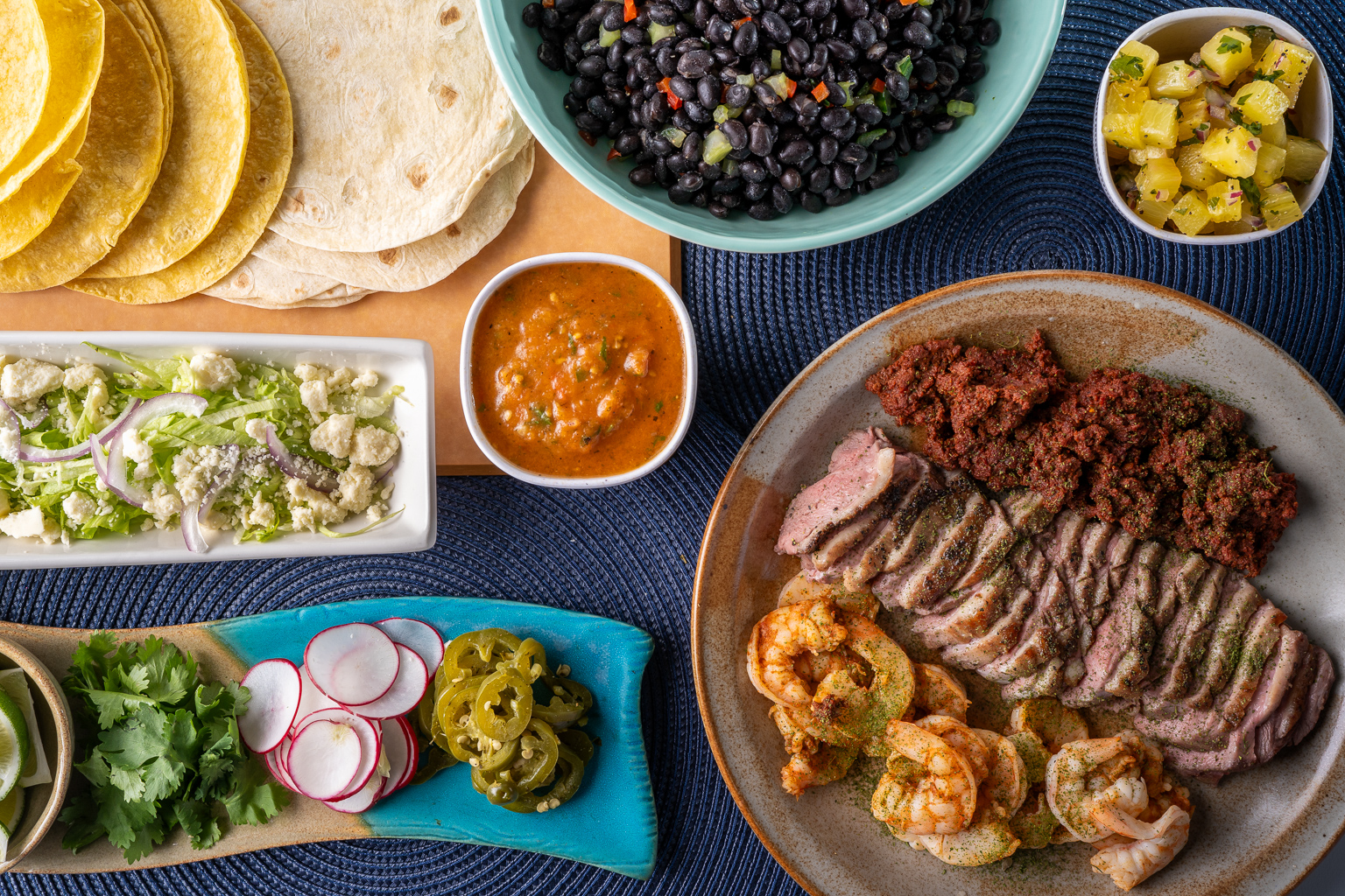A beautifully arranged platter featuring grilled steak, shrimp, and various side dishes including tortillas, black beans, fresh salad, salsa, and garnishes like cilantro and radishes.