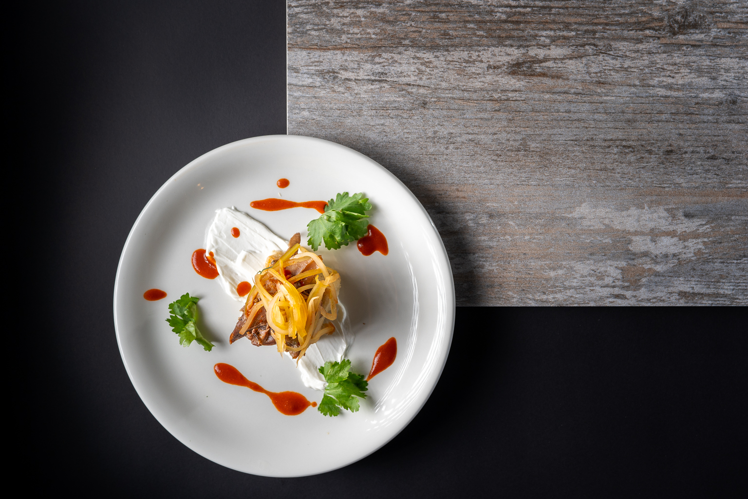 A beautifully plated dish featuring a savory meat component topped with yellow onions and fresh cilantro, drizzled with sauce on a white plate against a dark background.