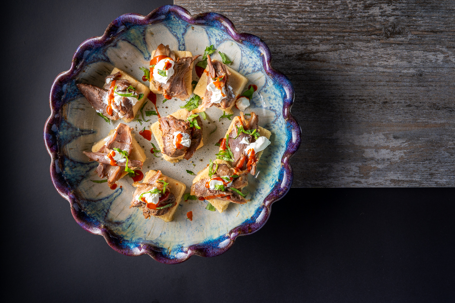 A beautifully arranged platter of bite-sized appetizers featuring pieces of seasoned meat on small squares of bread, garnished with creamy toppings and herbs, all served on a decorative dish.
