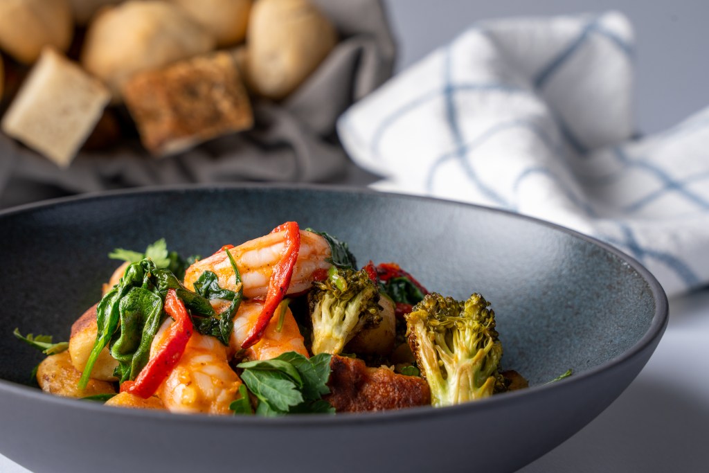 A beautifully plated dish featuring shrimp, sautéed vegetables, and herbs served in a dark bowl, with soft bread rolls in the background.